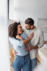 joyful multiethnic couple laughing while hugging at home