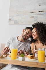 cheerful interracial couple smiling near breakfast on tray in bedroom