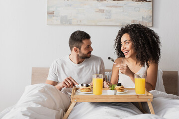 smiling african american woman holding fork with blackberry near boyfriend in bedroom