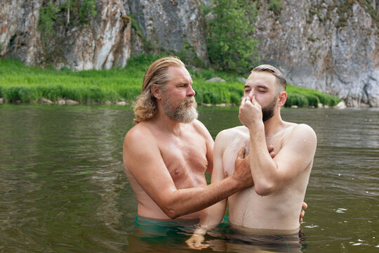 Two men in waist-deep water, young man with eyes closed and nose pinched by hand listens of older man. Ceremony of Christening outdoor
