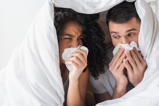 interracial sick couple under blanket sneezing in napkins