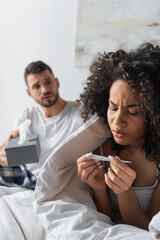 sick african american woman looking at digital thermometer near worried boyfriend on blurred background