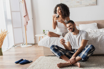 Fototapeta premium happy african american woman holding cup with coffee while bearded boyfriend using smartphone and sitting on carpet