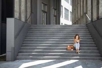 Pretty woman have outdoor lunch near office building while sitting on the stairs. Healthy meal for takeaway. With lowered protective mask on her chin and social distance alone. Copy space.
