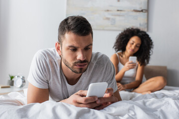 bearded man lying on bed and messaging on smartphone near african american woman on blurred background