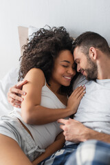 Fototapeta premium high angle view of cheerful interracial man and woman hugging while lying in bed