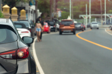 Cars parked in a queue beside the asphalt road. And blurry images of people walking and cars passing by in front.