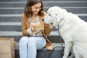 Pretty woman have outdoor lunch near office building with her big white dog while sitting on the stairs. Shares food with a dog. Pet friendly and pet care concept. Animal lover. 