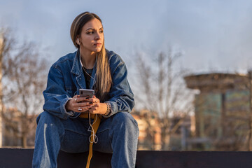 Obraz premium Nice young woman sitting on a bench in a public park with her smartphone