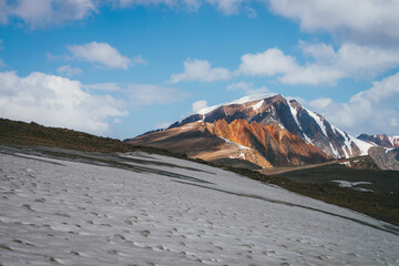 Scenic alpine landscape with snow-capped mountain peak and vivid red sharp rocks under blue sky with clouds. Colorful sunny mountain scenery with snow mountain top and pointy motley orange rocks.