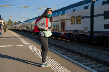 Asian backpacker girl traveling in times of covid19 - young happy and beautiful Japanese woman in face mask and backpack waiting for train at station platform visiting Europe