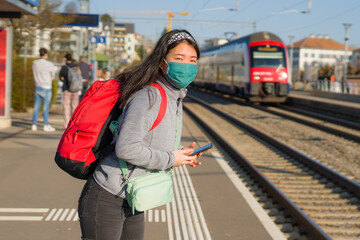 Asian backpacker girl traveling in times of covid19 - young happy and beautiful Korean woman in face mask and backpack waiting for train at station platform visiting Europe