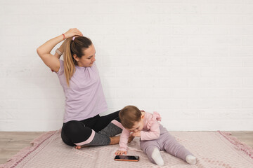 Fototapeta na wymiar young mother do sport exercises at home and her 1 year old daughter uses smartphone. multifunctional motherhood concept