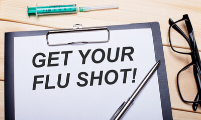The words GET YOUR FLU SHOT is written on a white piece of paper next to black-rimmed glasses, a pen and a syringe. Medical concept