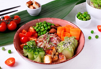 Vegan poke bowl with fried tofu, tomato, carrot, avocado, rice, cucumber, green pea and almond on the white table