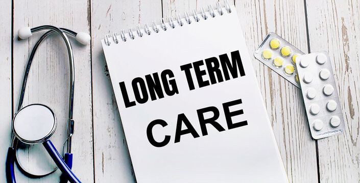 On a light wooden table lie a stethoscope, pills and a notebook with the inscription LONG TERM CARE. Medical concept