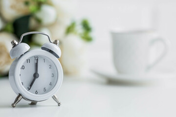 White retro alarm clock, flowers and a cup of coffee on the background of a white brick wall. The...