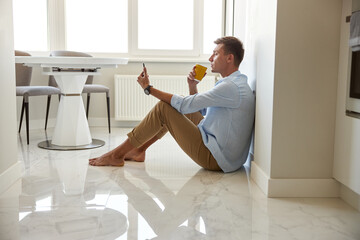 Caucasian hadsome young man in blue shirt is sitting on the floor and chilling at modern kitchen