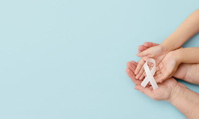 A white ribbon in the hands of a child and a mother or grandmother on a blue background.Concept lung cancer awareness November month. Top view. Copyspace