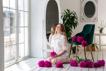 Woman in white dress sits on wooden floor among flowers of pink peonies, in white room. Portrait of blonde female. Surprise bouquet of flowers for date, mothers day or valentines day.