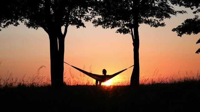 People on vacation. A girl's silhouette in a hammock between trees. A hammock in the background of the sunset. Rest and relaxation in nature. 