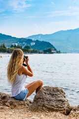 The girl sitting near water with photo camera. Blonde takes pictures of the mountains. Back view, vacation concept