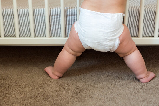Toddler attempting to sit down independently while holding onto crib rails; baby wearing diaper 