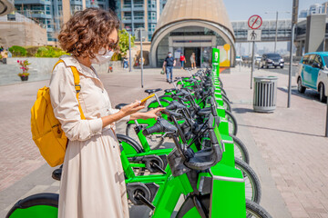 A woman wearing a medical mask uses a smartphone app to rent a shared electric bike to travel...