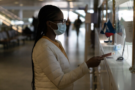 Black Woman Wear Protective Mask At Check-in Desk In Airport During Covid Epidemic. African Female Give Passport, Ticket For Registration At Terminal Counter Before Flight Under Corona Virus Disease