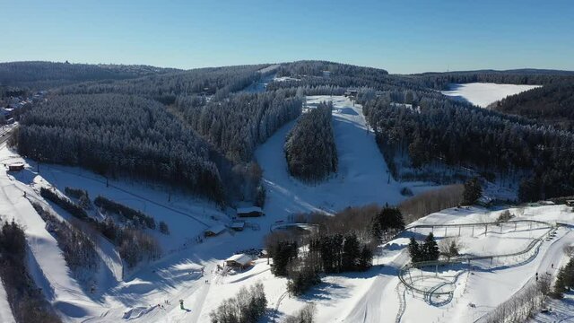 Air view on the winter sports slopes at the ski lift Karussell Winterberg. Toboggan slope and ski slopes between snow-covered spruce forests. Flight goes down.