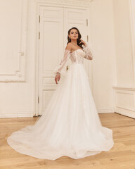 Fototapeta premium Elegant caucasian woman with natural make-up and hairstyle wearing white bridel dress, standing and posing in bright stylish interior. Full length portrait