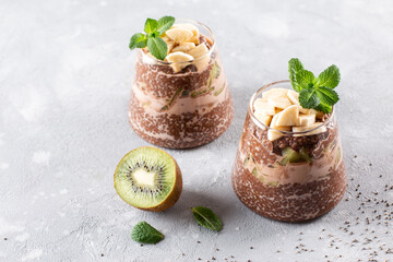 Chocolate chia pudding with kiwi, banana and mint on top in a glass jar on a gray concrete background. Copy space