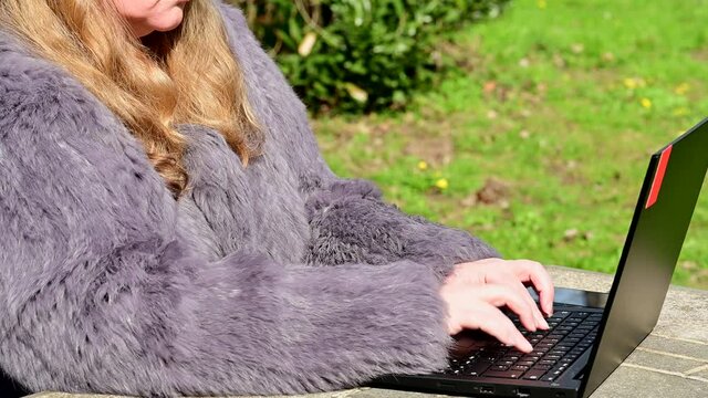 Frau in einer Kaninchenfell Jacke arbeitet im Garten mit einem Notebook im Homeoffice  