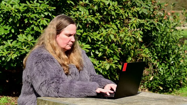 Frau in einer Kaninchenfell Jacke arbeitet im Garten mit einem Notebook im Homeoffice  