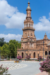 Fototapeta premium Spanish Square, in the center of Seville. Very touristic travel destination empty due to coronavirus measures. View of the south tower. Coloured flowers, blue sky with few clouds. 
