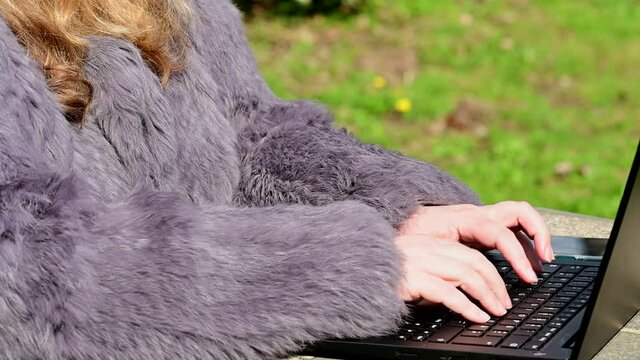 Frau in einer Kaninchenfell Jacke arbeitet im Garten mit einem Notebook im Homeoffice  