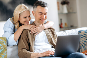 A middle aged married happy caucasian couple at home on the couch is using laptop. Joyful husband...