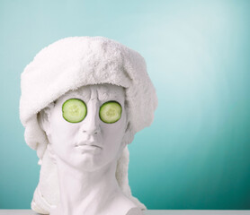 Plaster statue head blue background wearing white towel and cucumber face mask. Beauty care concept.