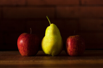 Two apples and a pear lined up on a wooden table with brick background.