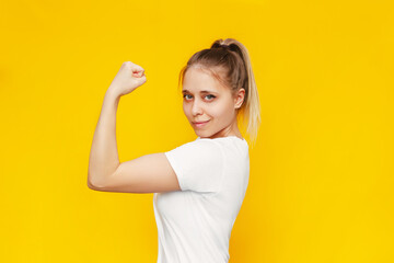 Strong powerful confident caucasian young blonde woman raises arm and shows bicep isolated on a bright colorful yellow background. Feminism, girl power, equal women's rights and independence concept