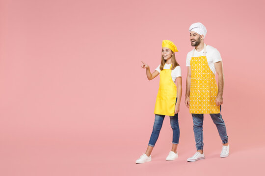 Full Length Body Of Teen Fun Girl Dad Man Father's Helper Chef Cook Confectioner Baker In Yellow Apron Toque Cap Uniform Walk Go Point Finger Aside Isolated On Pastel Pink Background Studio Portrait