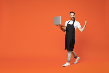 Fototapeta na wymiar Full length young man barista bartender barman employee in black apron white tshirt work in coffee shop using laptop pc computer do winner gesture isolated on orange background Small business startup