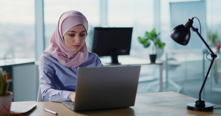 Young beautiufl arabic woman in business office wearing hijab working on laptop typing keyboard. Corporate girl. Traditional Muslim.