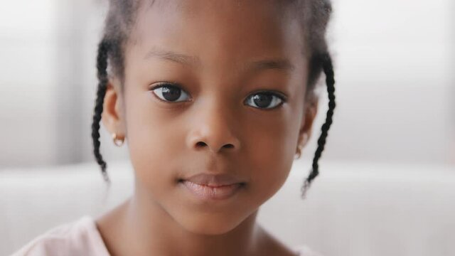 Close-up cute face african kid ethnic child looking at camera indoor, portrait of alone one little girl black daughter afro american toddler baby preschool schoolgirl with sad expression look at home