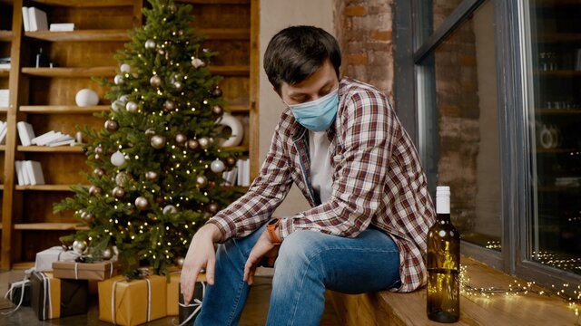 Problems Of Alcoholism In Loneliness, Sadness And Melancholy During Pandemic Covid 19. Male Removes Mask From Coronavirus, Drinking Wine From Bottle During New Year Holidays Sitting By Christmas Tree