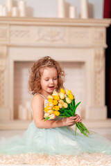 Portrait cute little girl in beautiful mint-colored dress with wavy hair and a tiara. baby is smiling happily and enjoying the flowers in the background of the fireplace, with a bouquet of tulips.