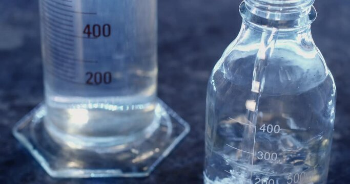 A clear liquid is poured into an empty flask and closed with a lid; the chemical glassware is illuminated with blue light.