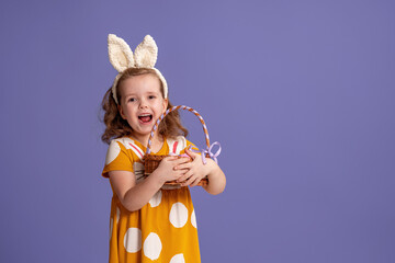 charming smiling little girl stands on a purple background in the studio and holds basket of Easter eggs. Funny happy baby wears rabbit ears on Easter day. Have fun hunting for Easter eggs. Copy space