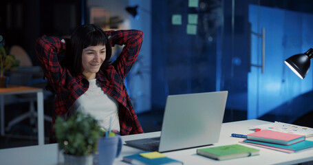 Joyful caucasian office woman doing computer project on laptop finishing work relaxing in chair...