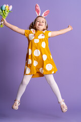 charming happy little girl with Easter bunny ears and a bouquet of spring tulip flowers made of fabric, jumping on a purple background. child smiles and rejoices at the arrival of spring and Easter